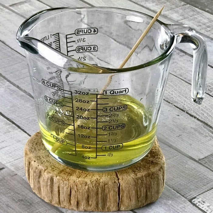 clear measuring cup on wooden riser with melted avocado butter and coconut oil and essential oils added to melt mix for Copycat Lush Ocean Salt scrub recipe made with essential oils
