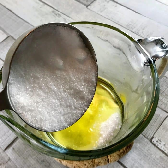 Adding salt to melted avocado butter and coconut oil in clear heat-safe measuring cup for Copycat Lush Ocean Salt scrub recipe made with essential oils