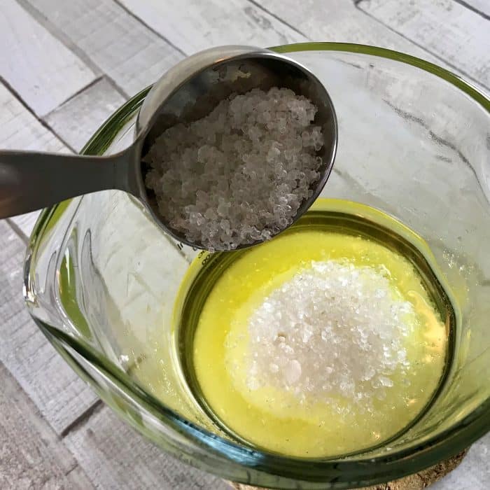 Adding coarse salt to clear heat-safe measuring cup containing sea salt and the melted mix of avocado butter and coconut oil for Copycat Lush Ocean Salt scrub recipe made with essential oils