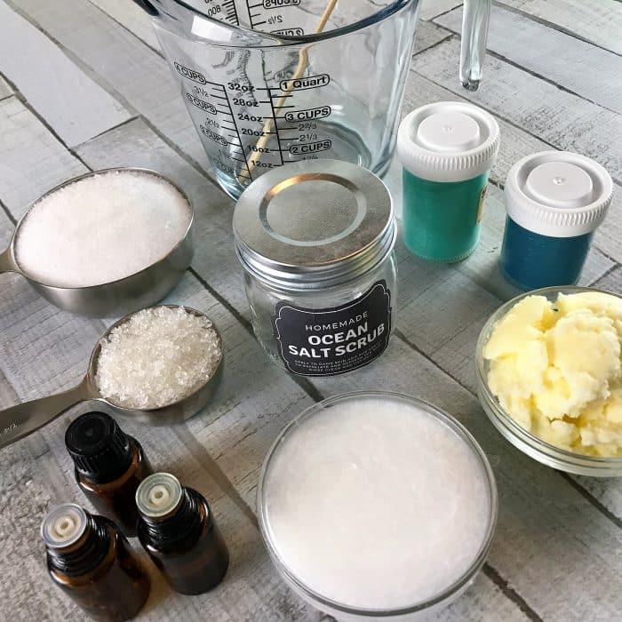 Ingredients display of Copycat Lush Ocean Salt scrub recipe made with essential oils: salts, avocado butter, coconut oil, mica, essential oils, plus equipment used in recipe measuring cups and clear glass jar