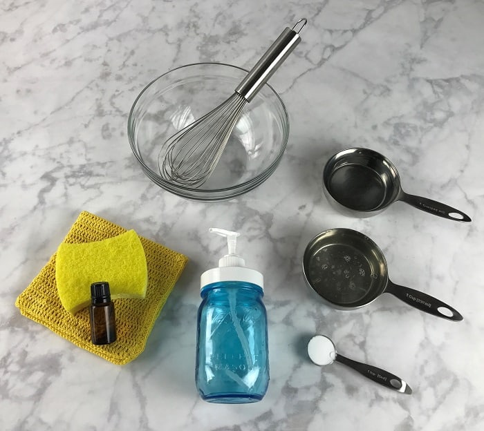 supplies to make homemade dish soap - mixing bowl, whisk, mason jar soap dispenser, essential oil, measuring cups with liquid, powder in measuring spoon, dish cloth, and sponge