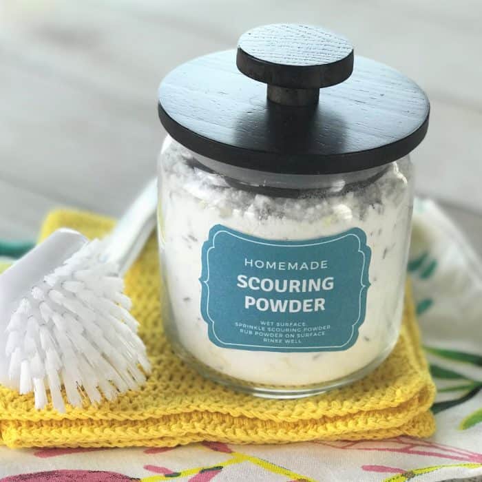 homemade scouring powder in glass canister with scrub brush beside it, on top of dish cloth and towel