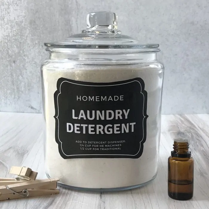 finished homemade laundry soap in a glass canister with lid, a free printable label has been added to the front of the jar and an essential oil bottle and clothes pins are on either side of the canister