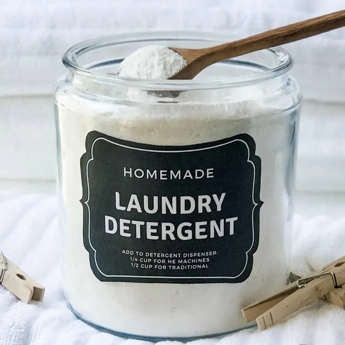wooden spoon scooping out powder laundry soap from large glass cansiter, clothes pins and fluffy white towels surround the laundry detergent canister