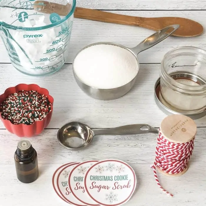 Sugar Scrub ingredients and tools display of coconut oil in measuring cup, organic non-gmo sugar in measuring spoon, red and green nonpareil sprinkles in cup, vanilla oleoresin in dropper bottle, wooden spoon, and custom lid labels