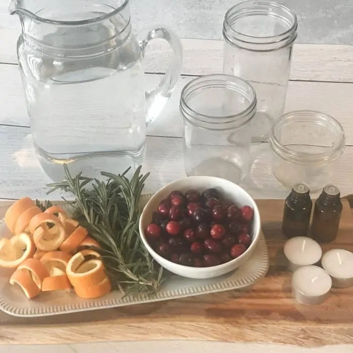 orange peels, rosemary sprigs, cranberries, essential oil bottles, pitcher of water, clear glass jars, essential oil bottles, ingredients for scented floated candles with essential oils