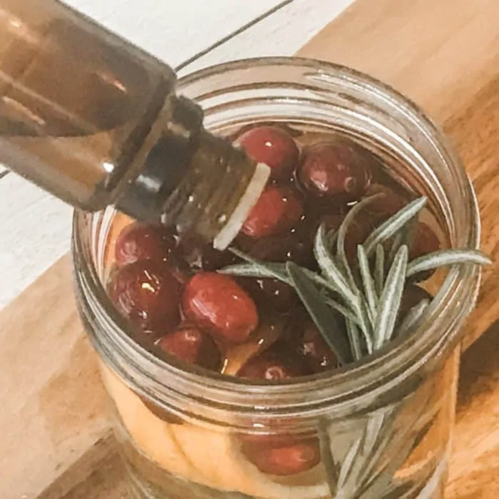adding essential oil to mason jar with cranberries, orange peels, rosemary sprigs, water, floating scented candle with essential oils