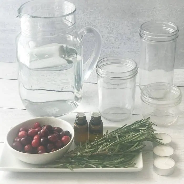 rosemary sprigs, cranberries, essential oil bottles, pitcher of water, clear glass jars, essential oil bottles, ingredients for scented floated candles with essential oils