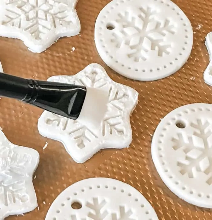homemade salt dough ornaments in snowflake shapes on cookie sheet, using brush to clean them