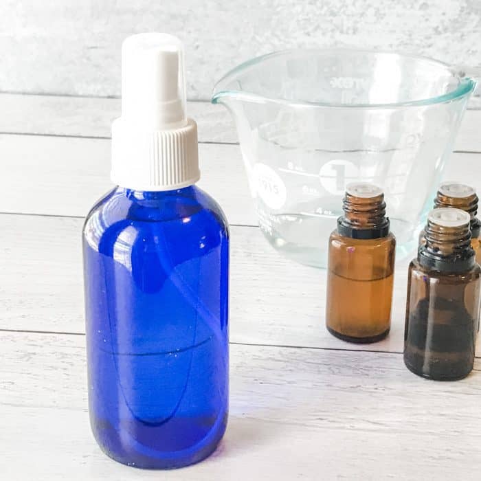 filled blue spray bottle in foreground, essential oil bottles and measuring cup in background, items used to make Winter Blues body spray with essential oils