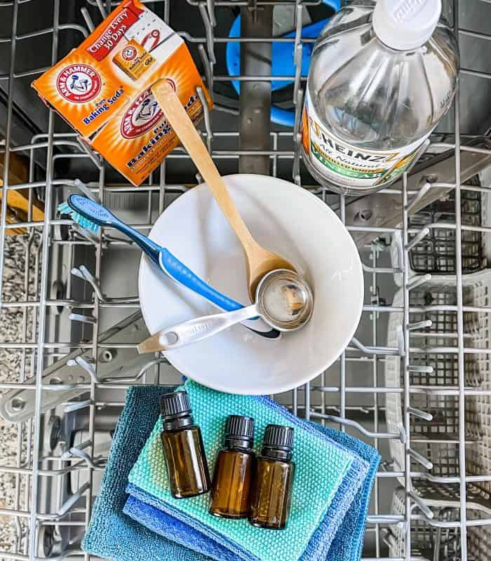 supplies for cleaning dishwasher (essential oil, baking soda, vinegar, cloths, toothbrush, bowl, and spoons)