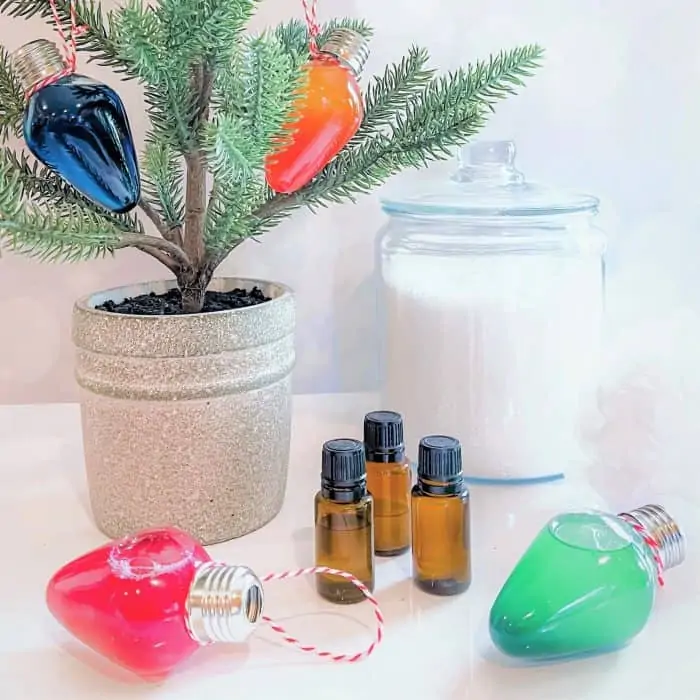 Christmas tree with orange and blue bath oil ornaments hanging from the branches -- red and green ornaments are on the table next to 3 bottles of essential oil and jar of bath salts