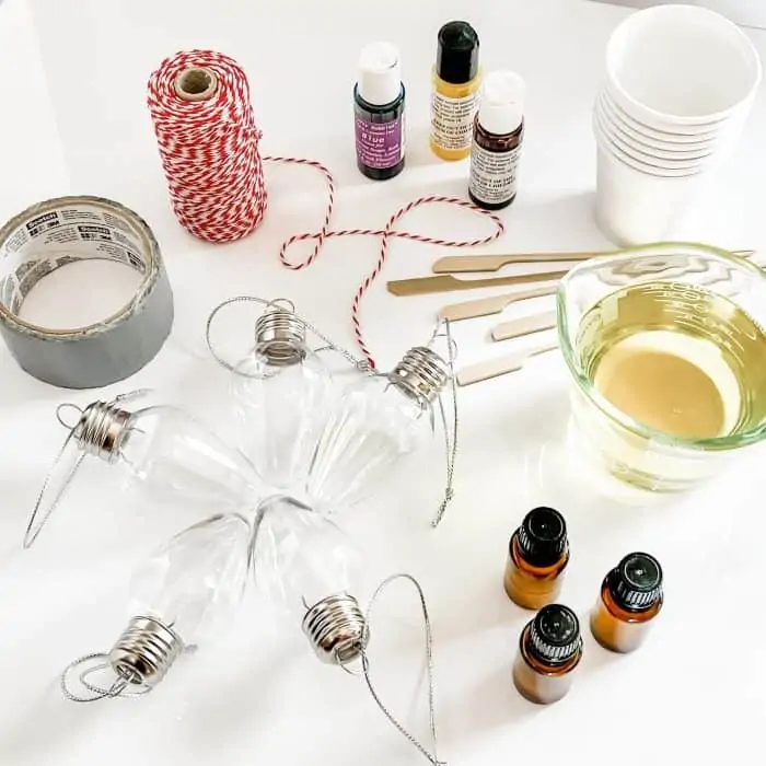 supplies to make homemade scented bath oil -- bath oil in measuring cup, 3 bottles of essential oil, clear Christmas lightbulb ornaments, duct tape, red & white twine, wood stirrers, soap colorants, paper cups