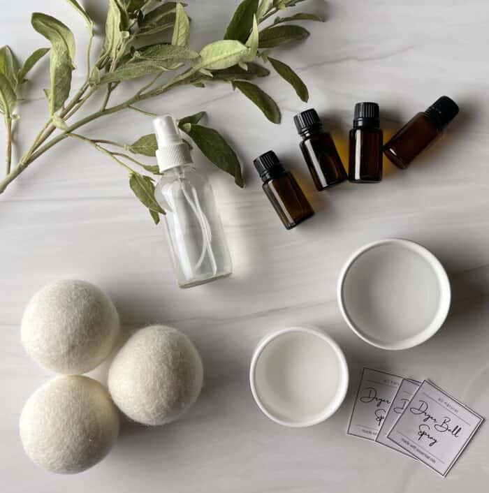 supplies to make DIY dryer ball spray - witch hazel, distilled water, essential oils, 4 oz glass spray bottle, free prittable dryer ball spray labels-- next to 3 wool dryer balls and greenery branch