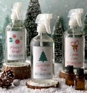 3 glass spray bottles of homemade all-purpose cleaner in favorite Christmas scents- CHristmas Tree, Candy Cane, and Merry & Bright. 2 essential oil bottles, a couple pinecones and CHristmas trees in background