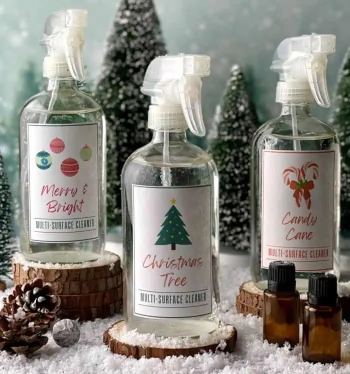3 spray bottles of DIY multi-purpose cleaner - Merry & Bright, Christmas Tree, Candy Cane. by oneessentialcommunity.com... next to 2 essential oil bottles, a few pinecones, and evergreen trees in background