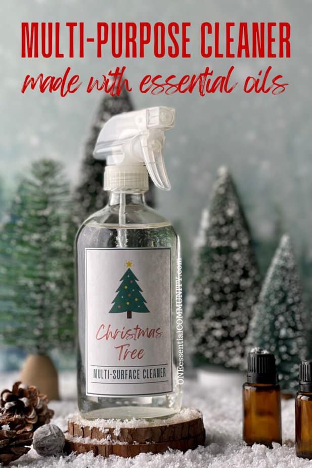multi-purpose cleaner made with essential oils by oneessentialcommunity.com -- glass spray bottle of Christmas Tree multi-purpose cleaner on top of a couple of wood slices next to two essential oil bottles and a few pinecones, background of evergreen trees 